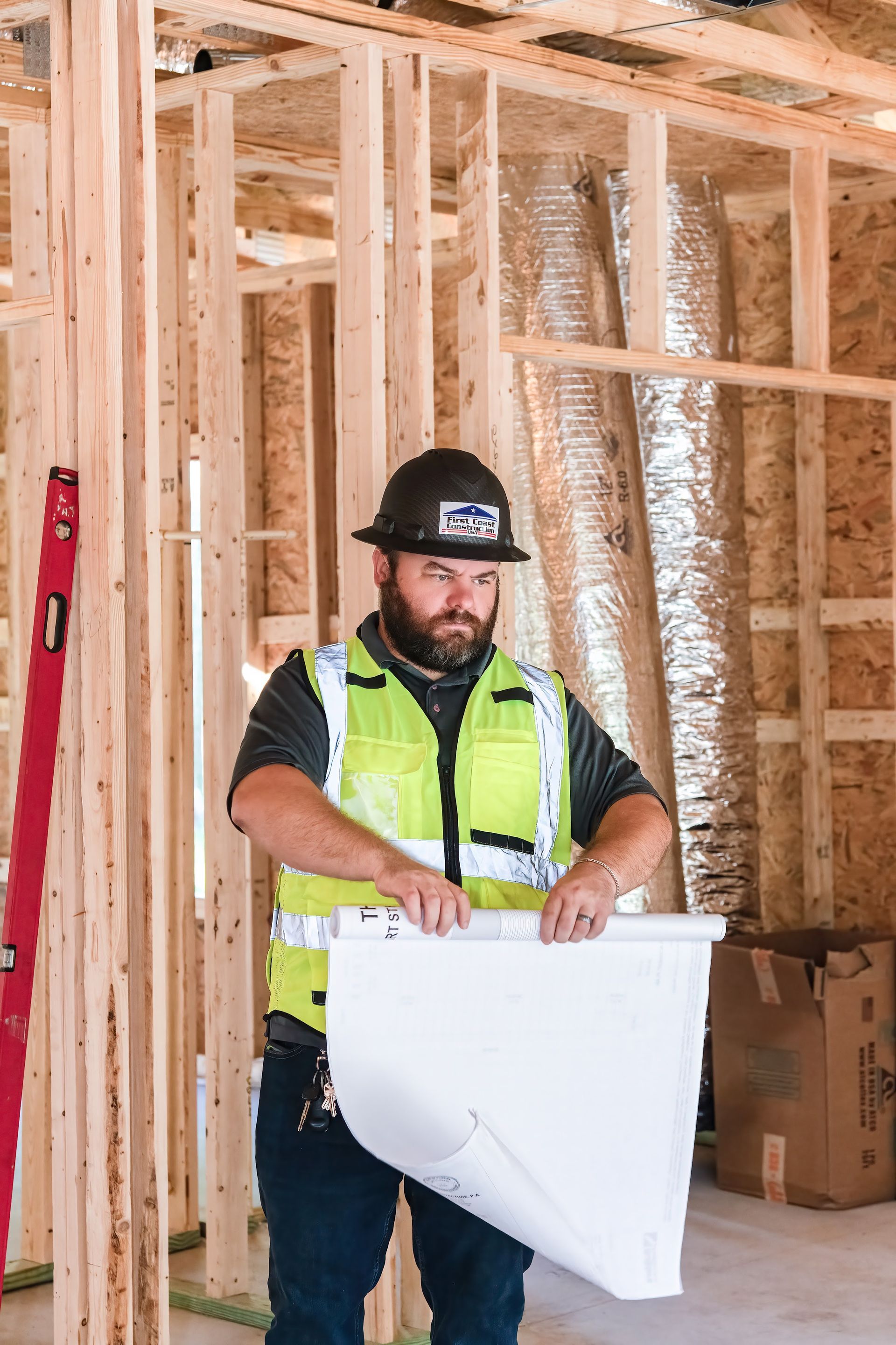 Construction worker in a hard hat and safety vest holding a blueprint inside a framed building site