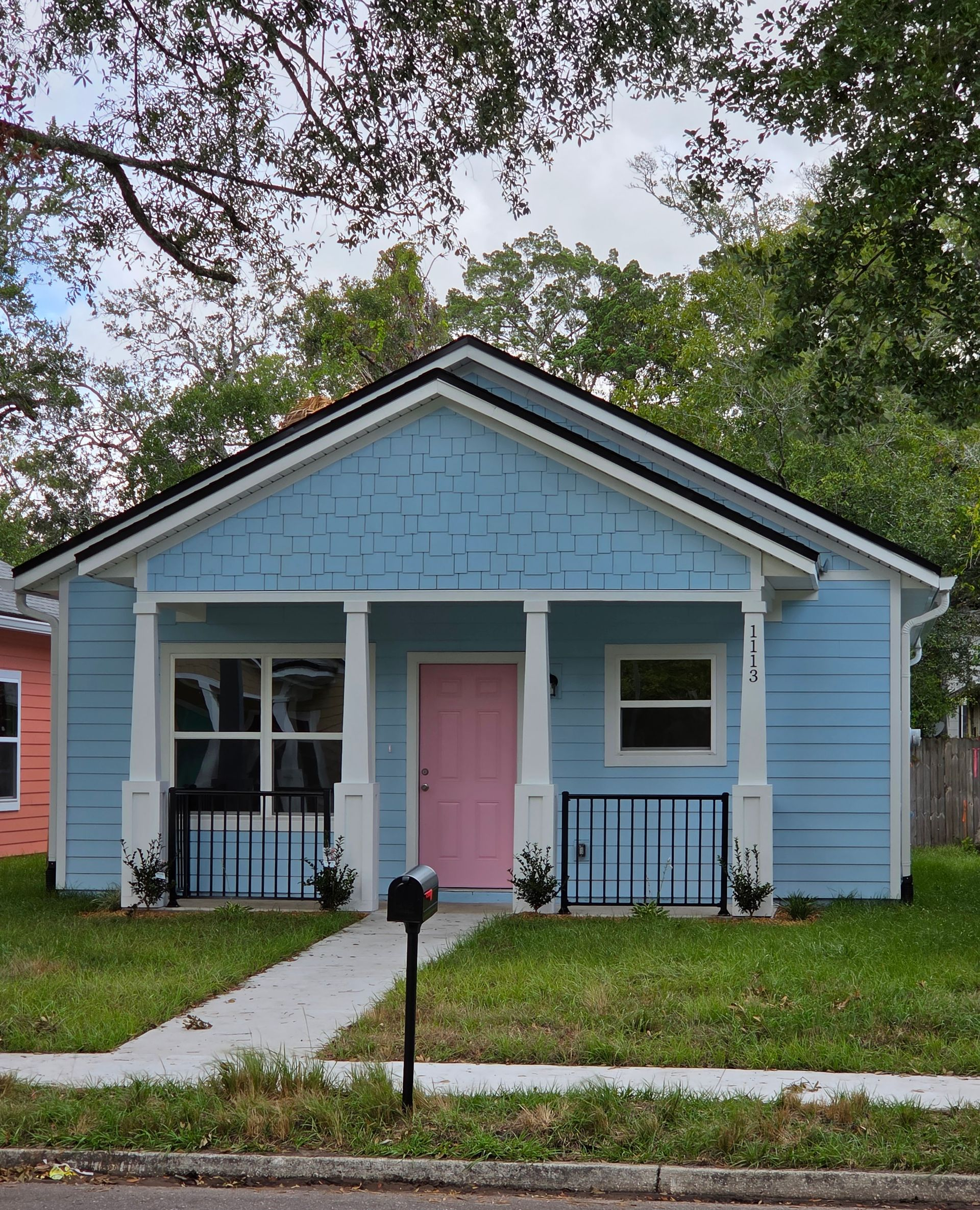 Small blue cottage with a pink front door, porch railings, and a front lawn under trees.