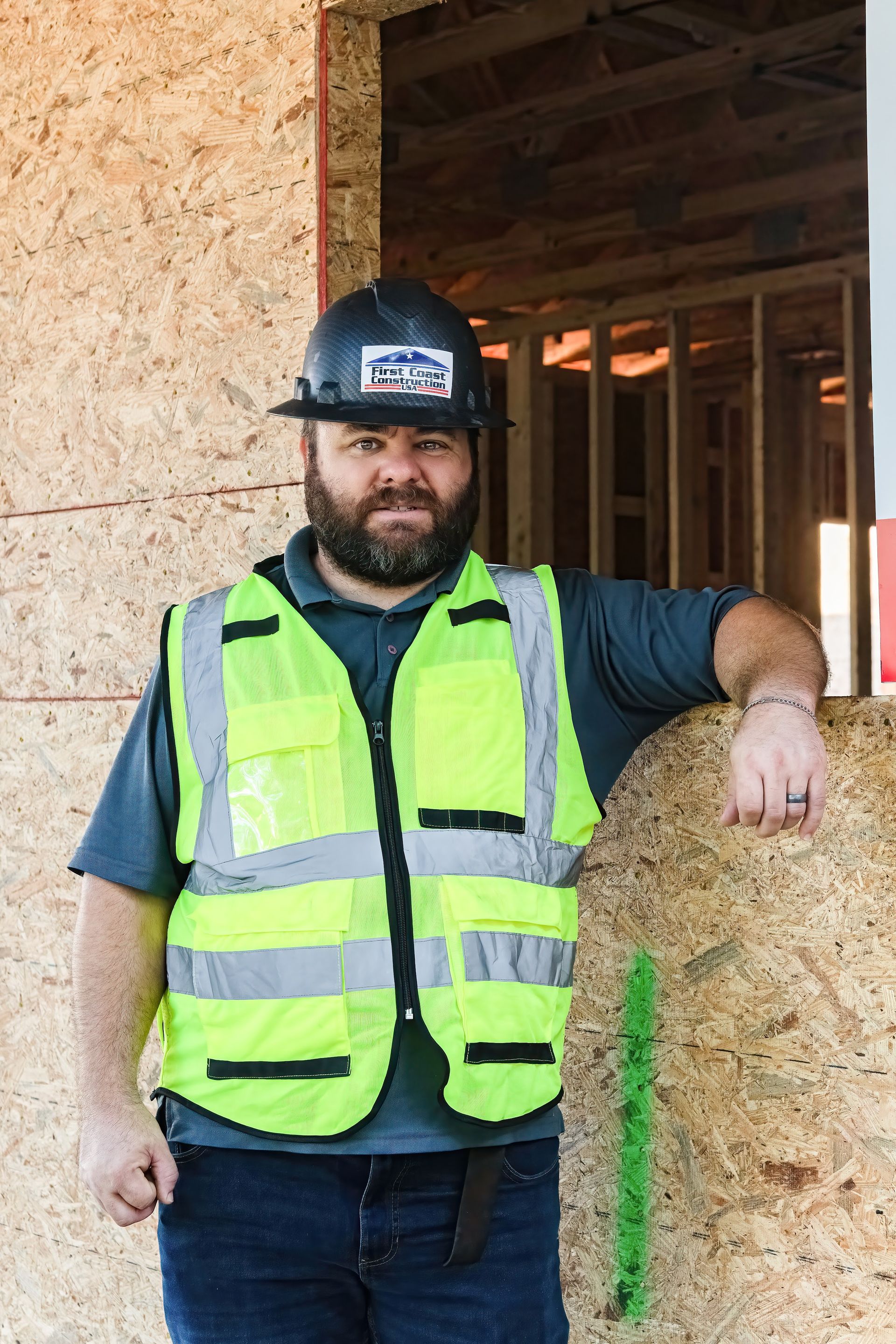 Construction worker in hard hat and neon vest leaning on a wooden frame at a building site.