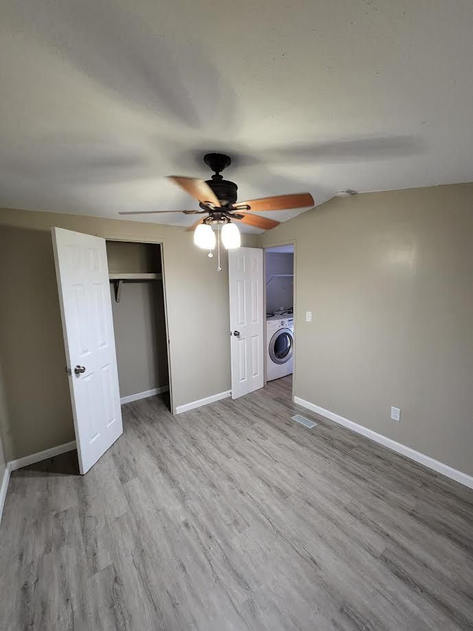 Empty beige bedroom with ceiling fan, open closet, and laundry area visible through doorway.