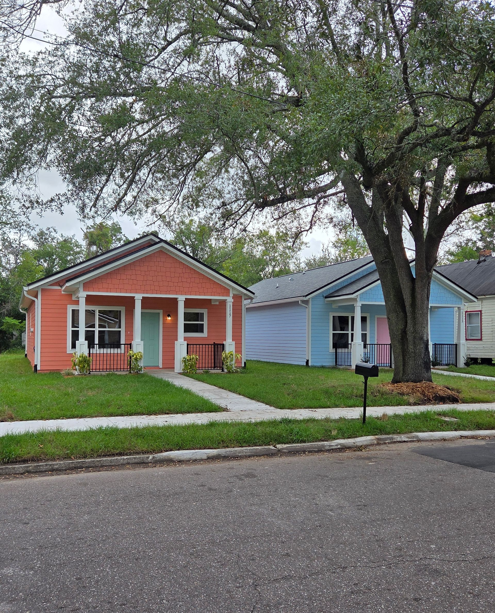 Two small bungalows, one red and one blue, with a front lawn and large tree along a quiet street.