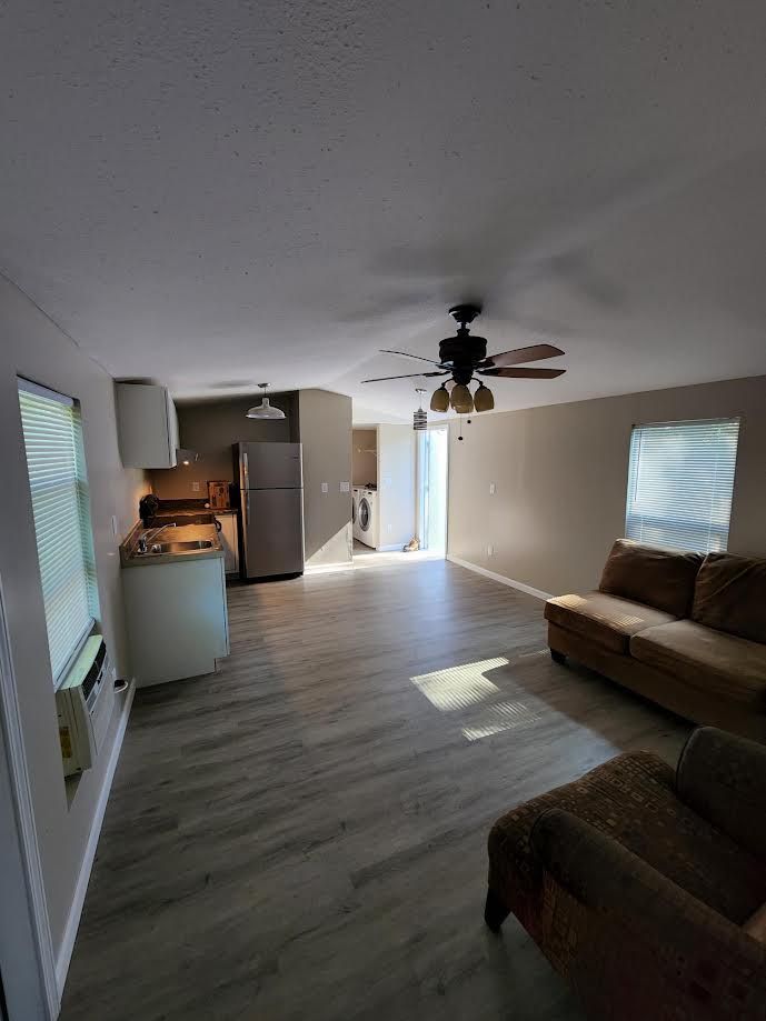 Bright open living room with wood floors, couch, ceiling fan, kitchen area, and windows with blinds
