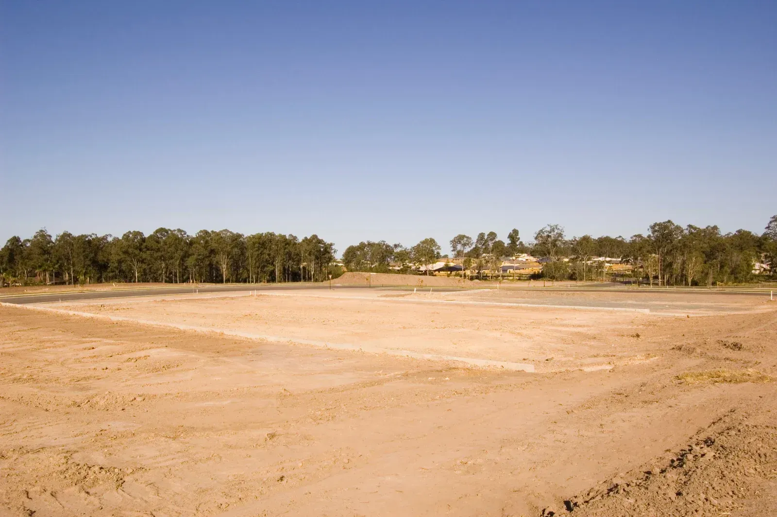 Wide barren sandy field under a clear blue sky, with a line of trees in the distance