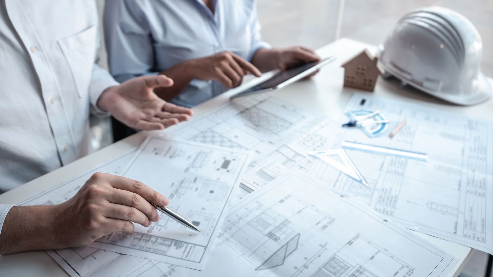 Engineers reviewing architectural blueprints at a desk with a white hard hat and tablet