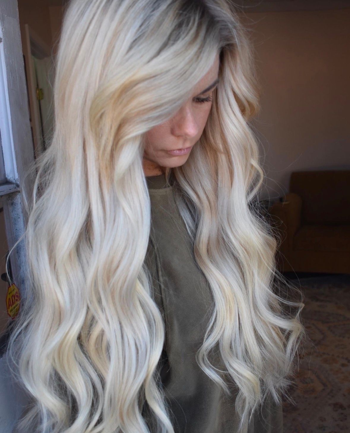 Person with long wavy platinum-blonde hair looking down indoors, wearing a gray top.