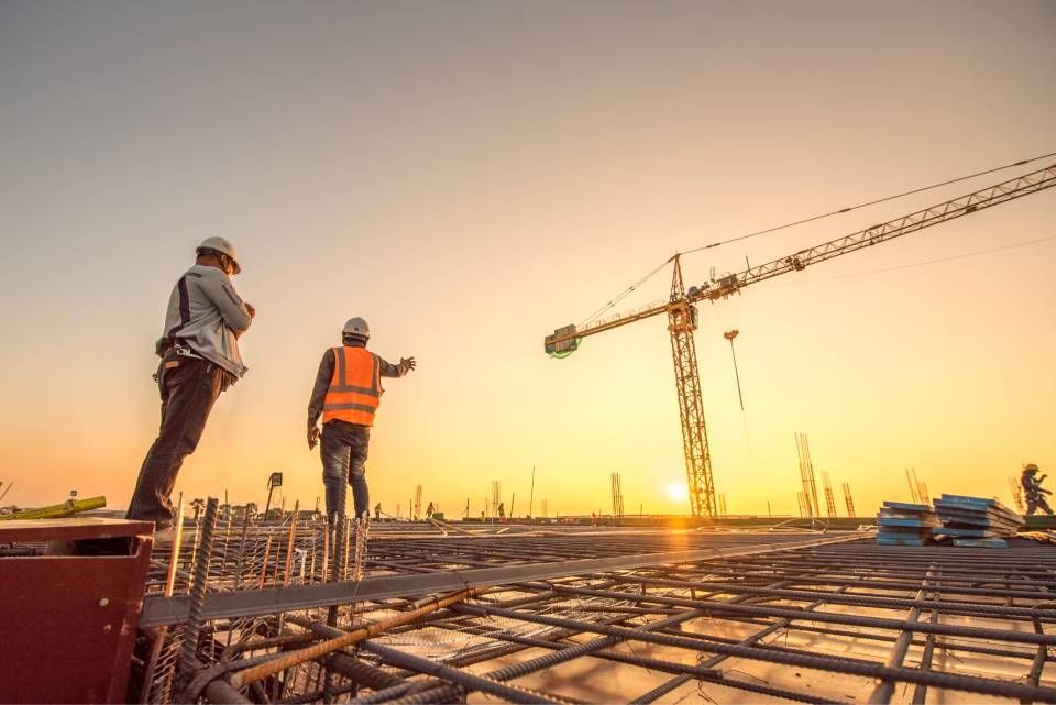Two workers in safety gear oversee a large construction site at sunset, with a tall crane in the background.