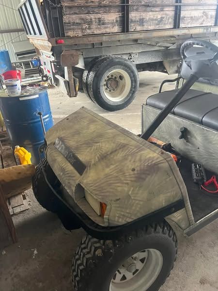 Camouflaged golf cart parked in a garage beside a pickup truck and blue barrel