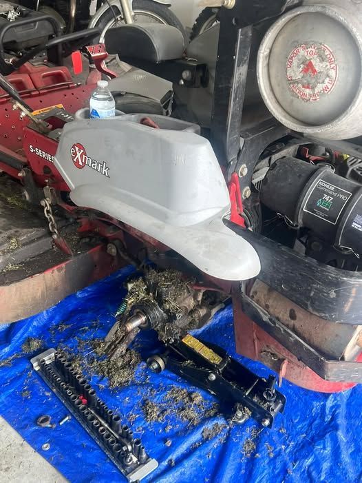 Red snowblower with gray cover on blue tarp inside a cluttered garage, parts removed and scattered nearby