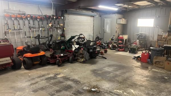 Cluttered workshop garage with lawn mowers, tools, and equipment along concrete walls and floor