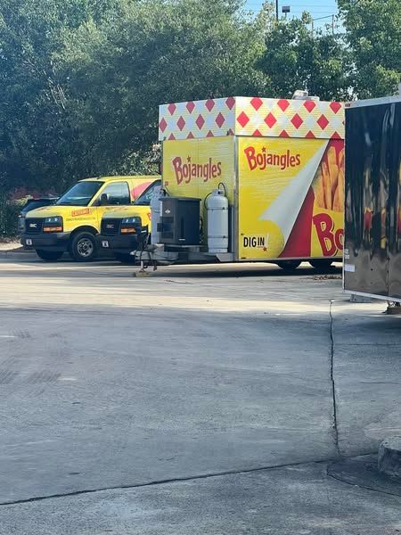 Bright yellow BJ’s truck and trailer parked by trees in a lot, with a dark vehicle beside it.