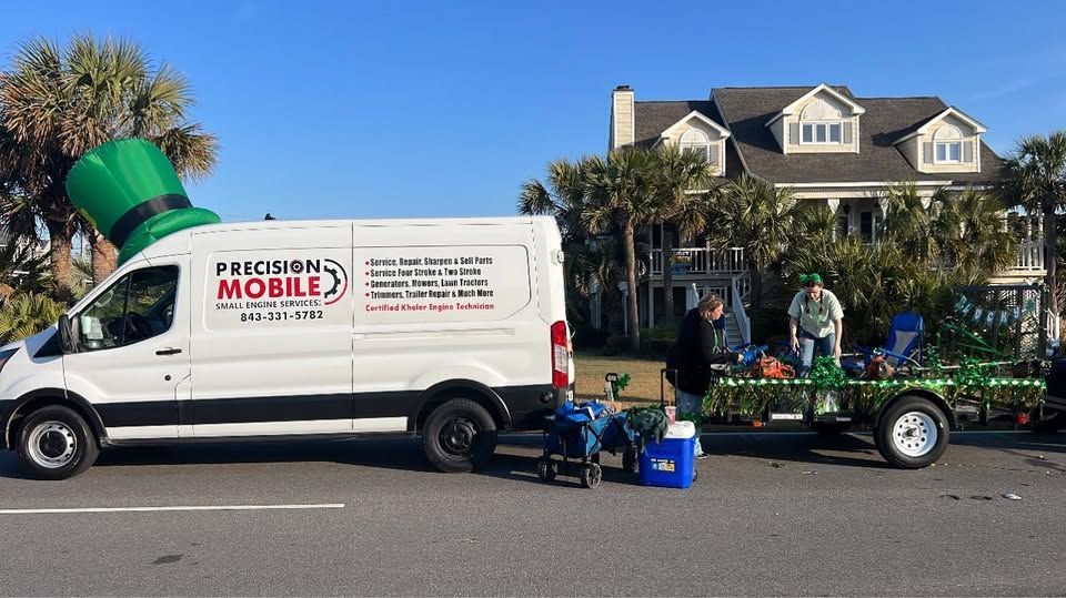 White mobile landscaping truck with large green blower parked on a street beside a trailer and workers.
