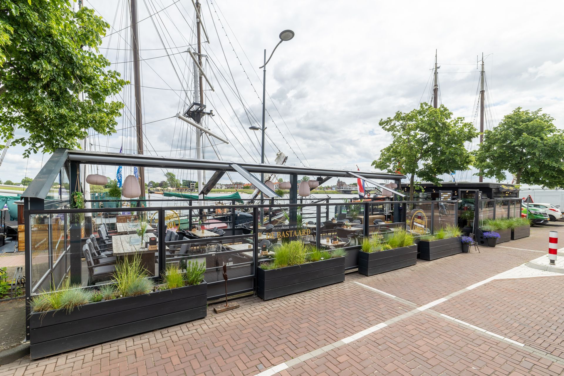 Terrasjes aan het water, gelegen aan een kanaal, met zwarte plantenbakken en aangemeerde boten op de achtergrond.