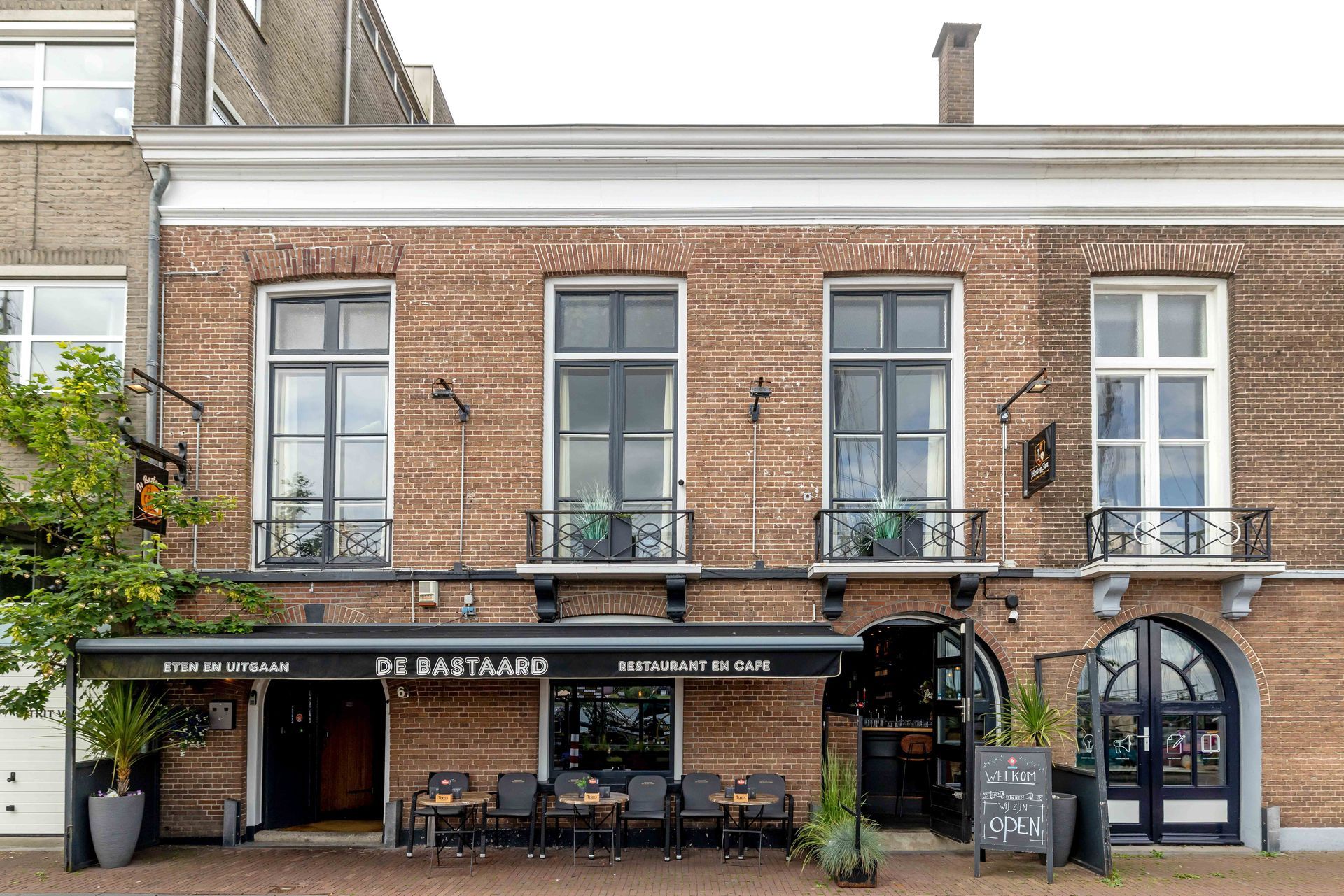 Een café met een bakstenen gevel, een terras en een zwarte luifel aan een stadsstraat. IJsselkade