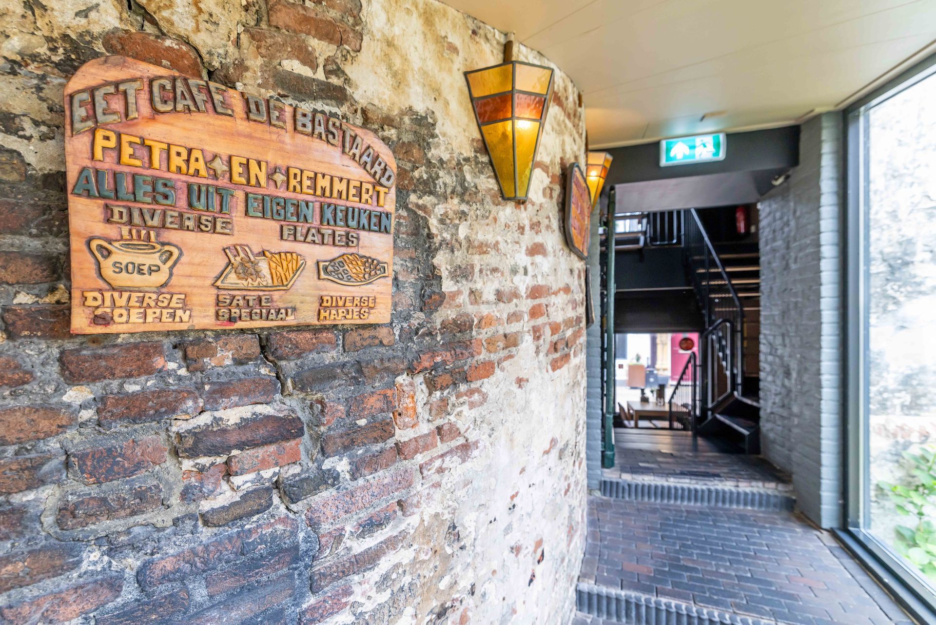 Bakstenen hal met vintage bierreclamebord, wandlamp en trap.