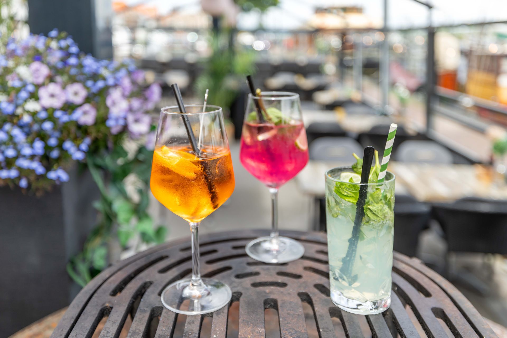 Drie kleurrijke cocktails op een ronde buitentafel met bloemen op de achtergrond.
Geniet hier van U drankje