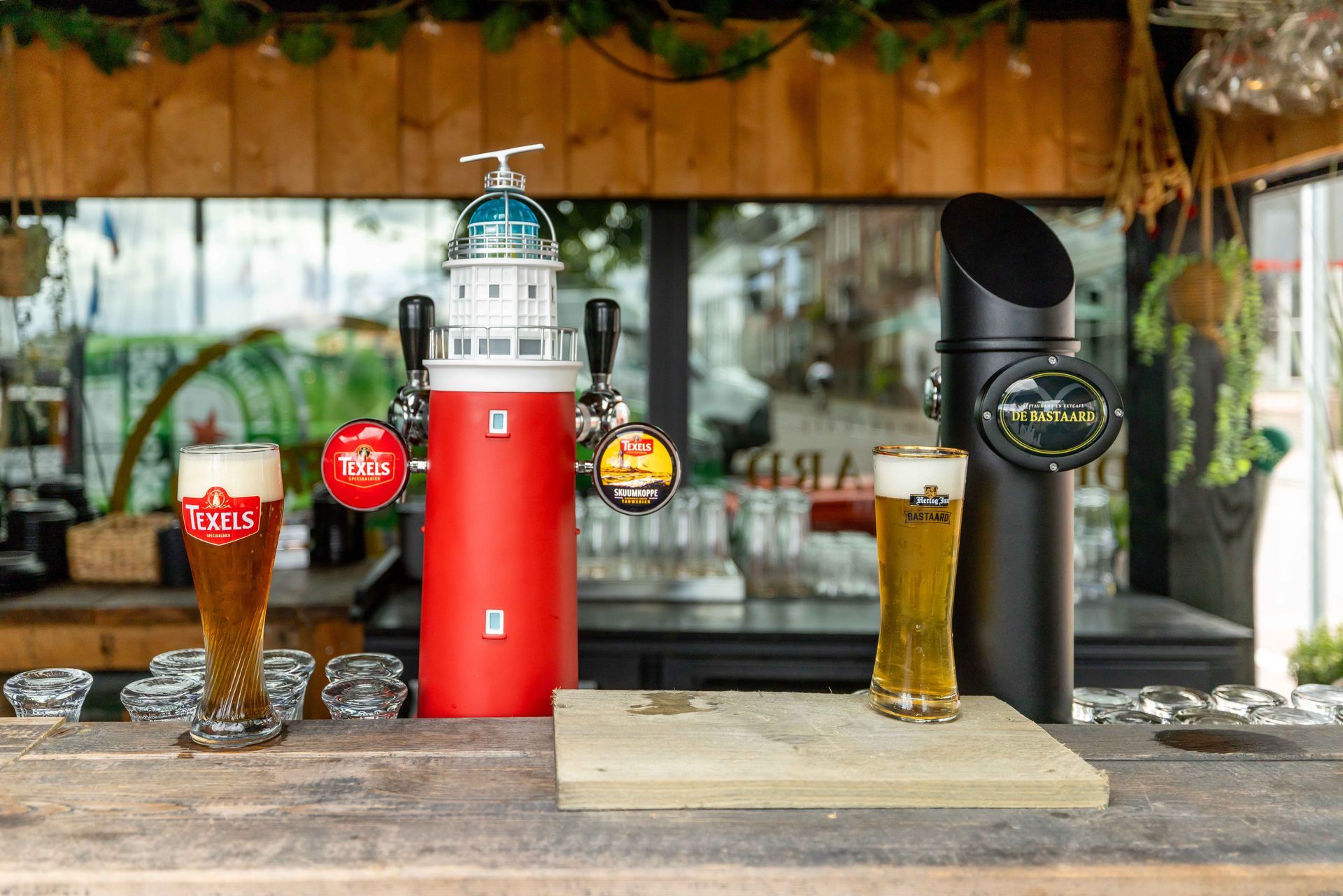 Twee biertjes naast een rode tapkraan in vuurtorenstijl aan een buitenbar.