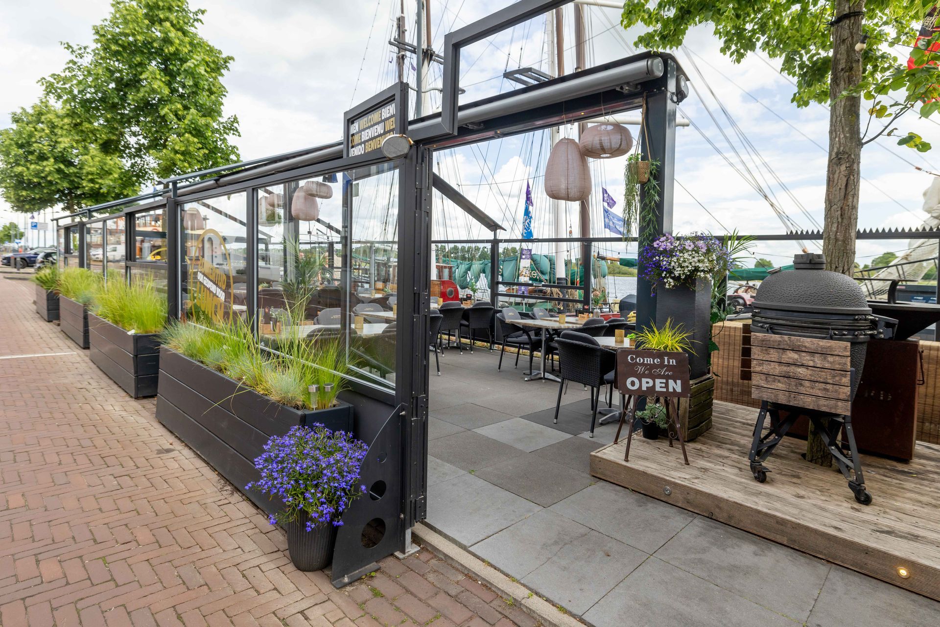 Een terras met een glazen omlijsting in zwart, een bakstenen pad en potplanten langs de stoep.