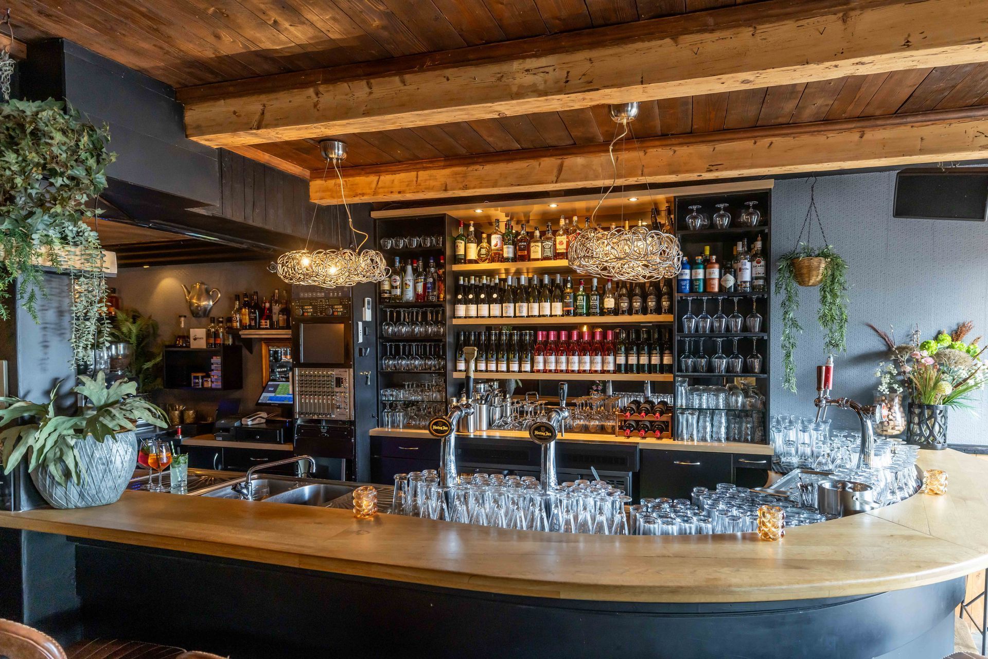 Gezellige bar met een gebogen houten toonbank, planken vol flessen sterke drank en een warm plafond met houten balken.