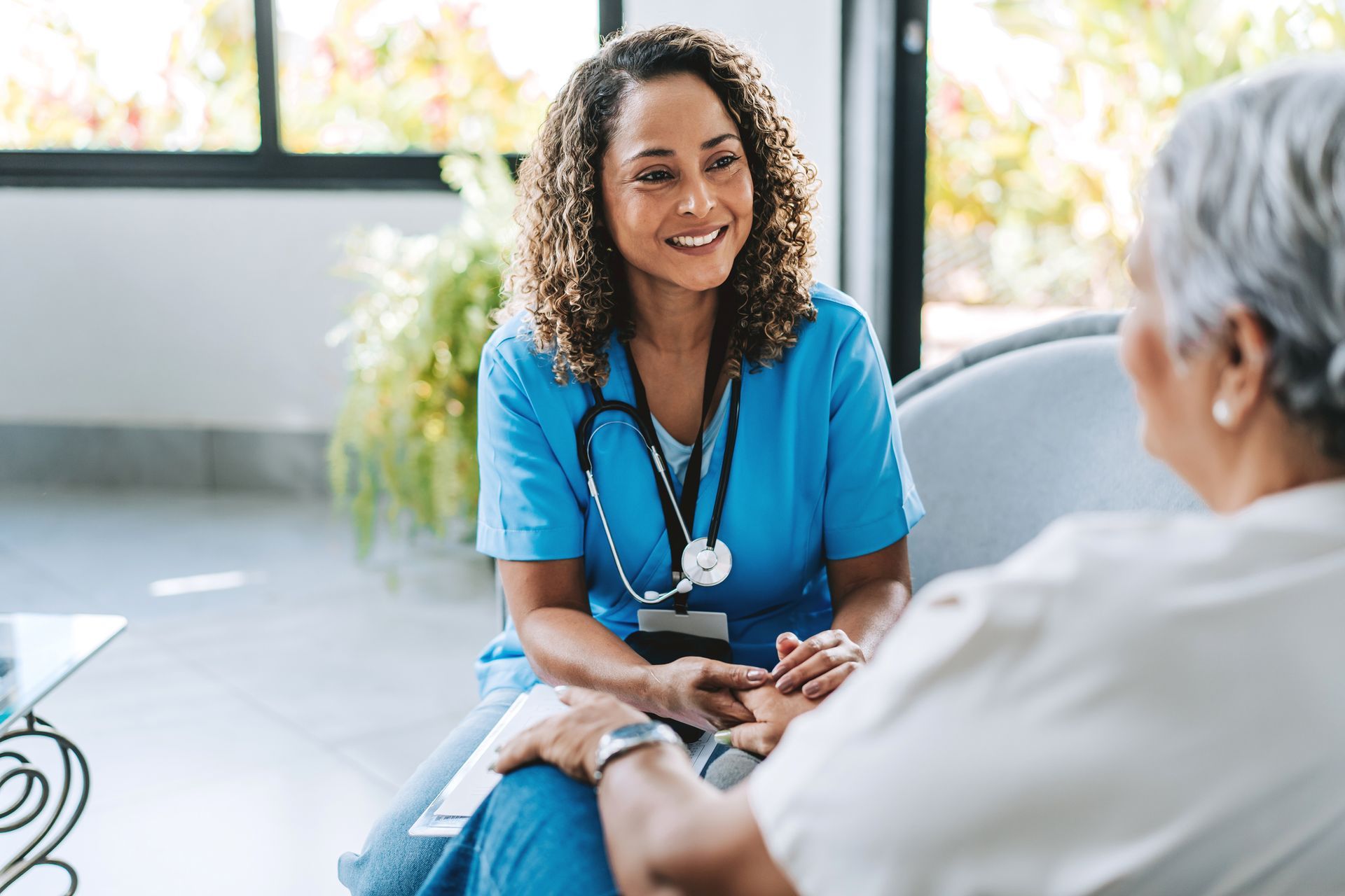 Nurse in blue scrubs smiling, holding the hand of an older woman.
