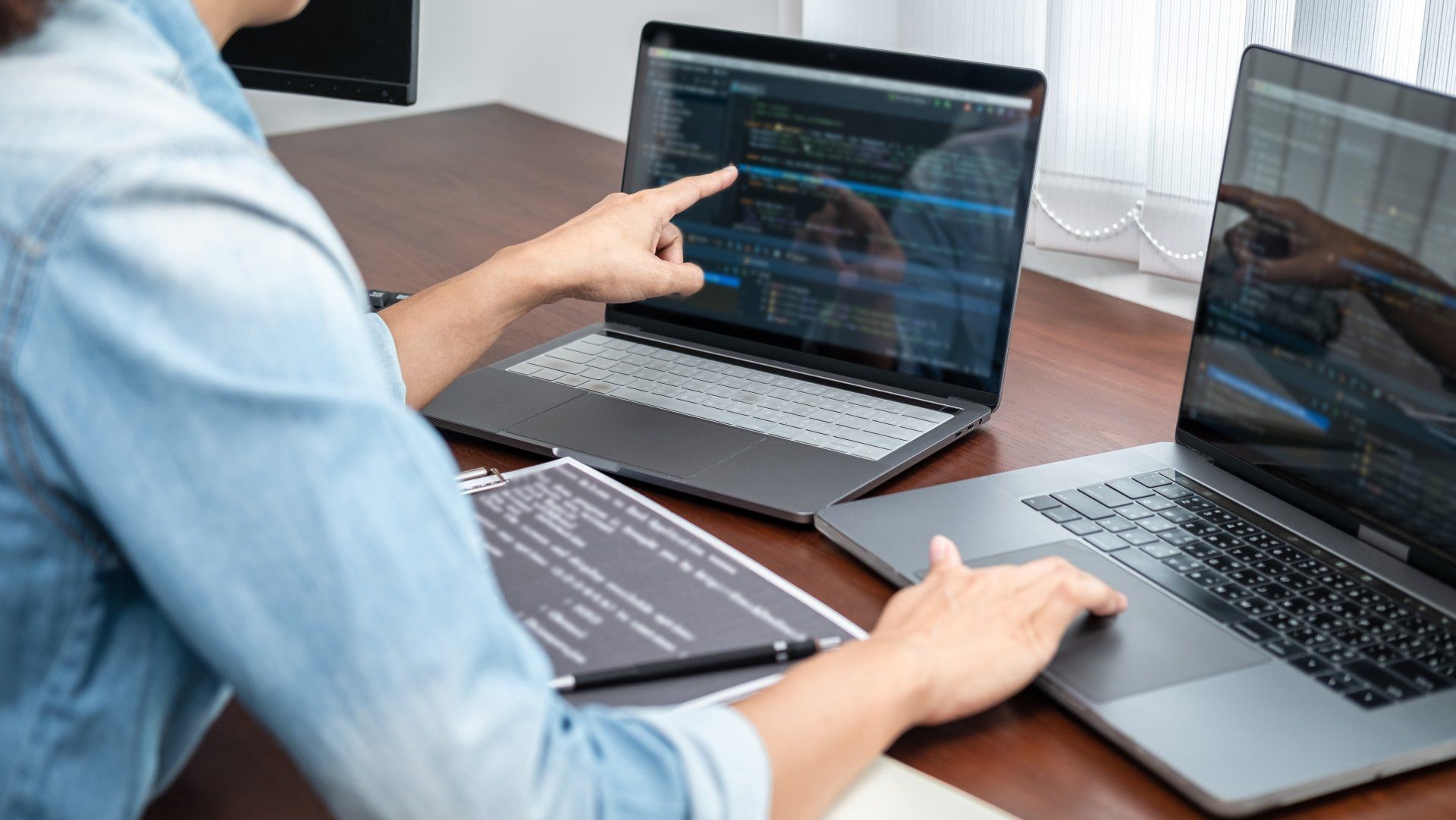 Person pointing at data on a laptop beside another laptop on a desk, with a notebook nearby.