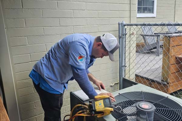 Technician in gray hoodie inspecting an outdoor HVAC unit beside a brick wall and chain-link fence