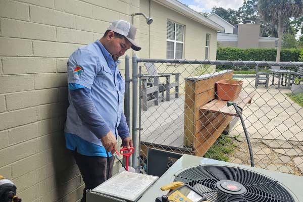 Technician in blue shirt servicing an outdoor HVAC unit beside a building.