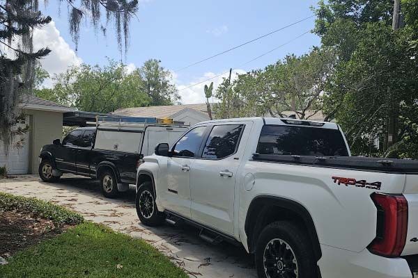 Two parked pickup trucks on a driveway beside a house, one white and one black, under palm trees.