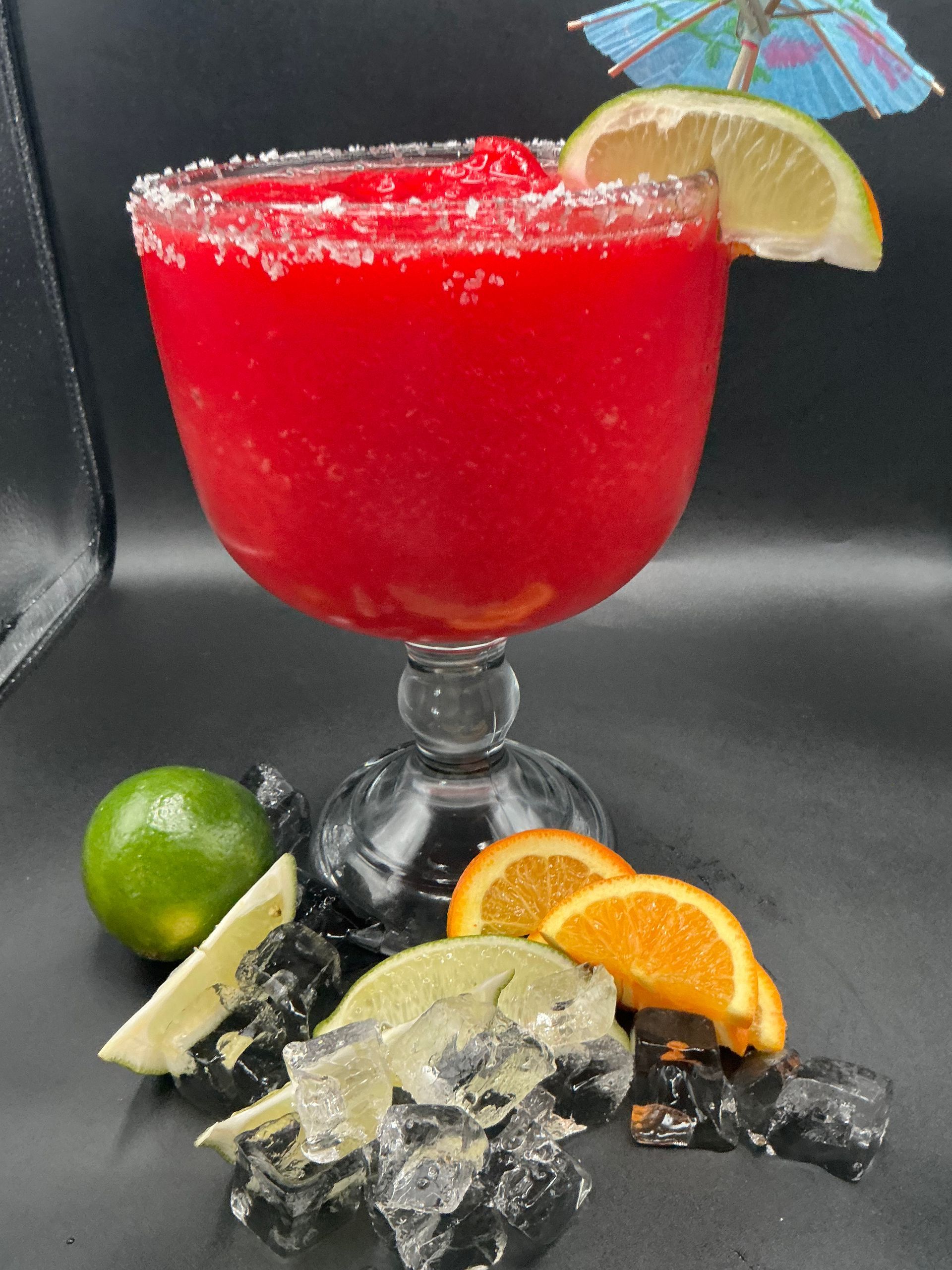 Red frozen cocktail in a salt-rimmed glass, garnished with lime and umbrella. Surrounded by fruit slices and ice.