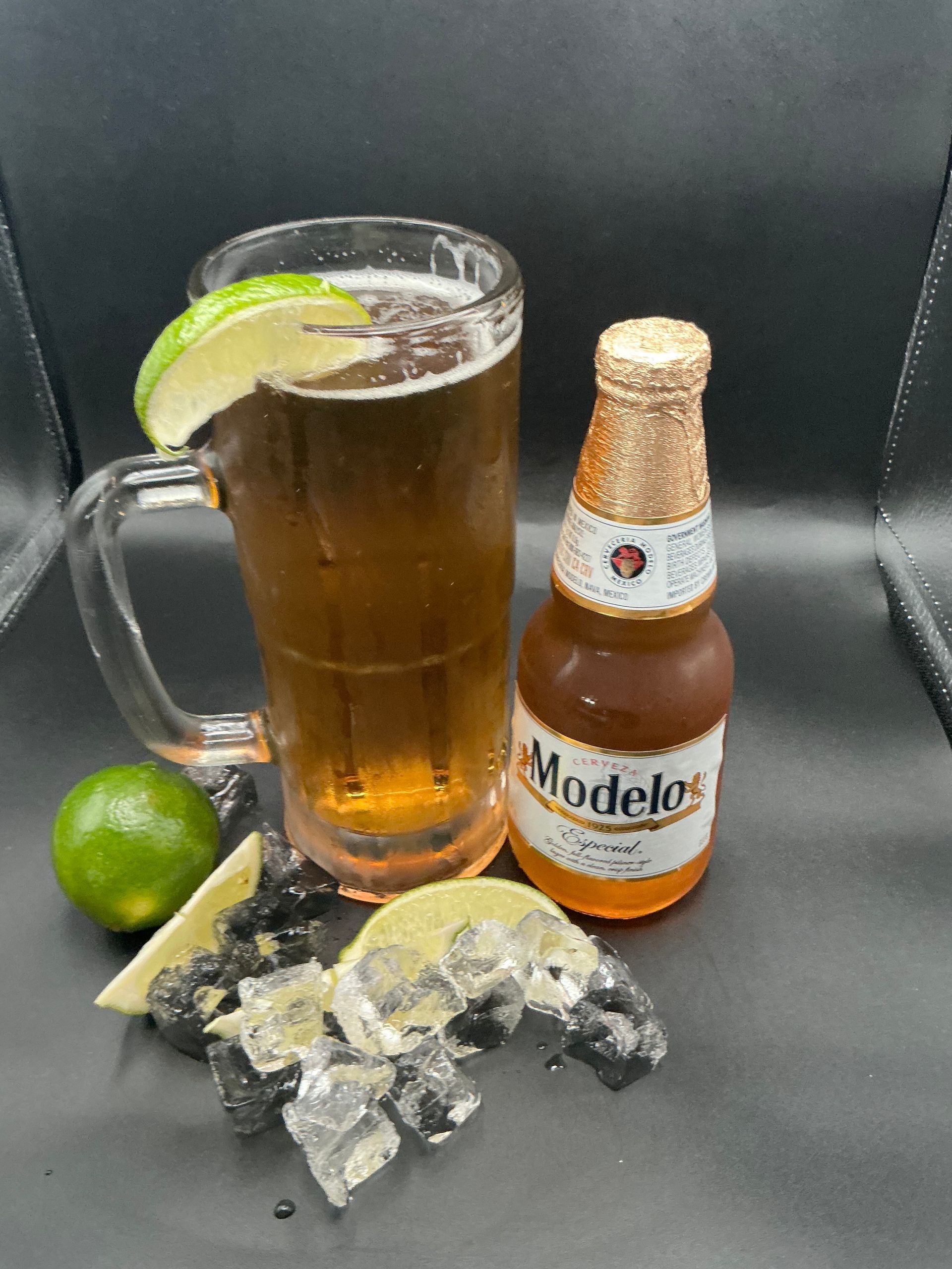 Mug of beer and Modelo bottle with lime wedges and ice on a black surface.