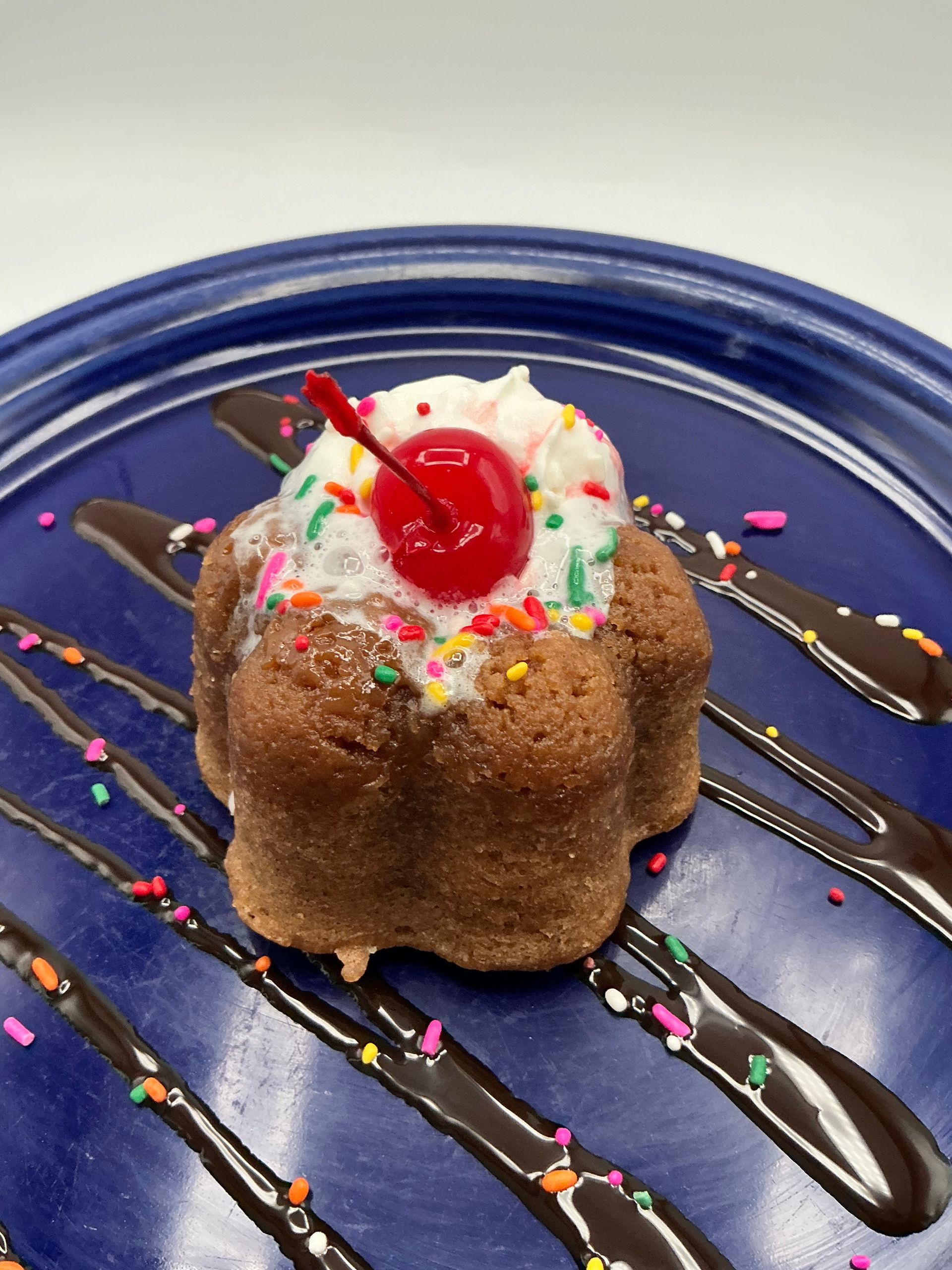 Mini cake with whipped cream, sprinkles, cherry, and chocolate drizzle on blue plate.