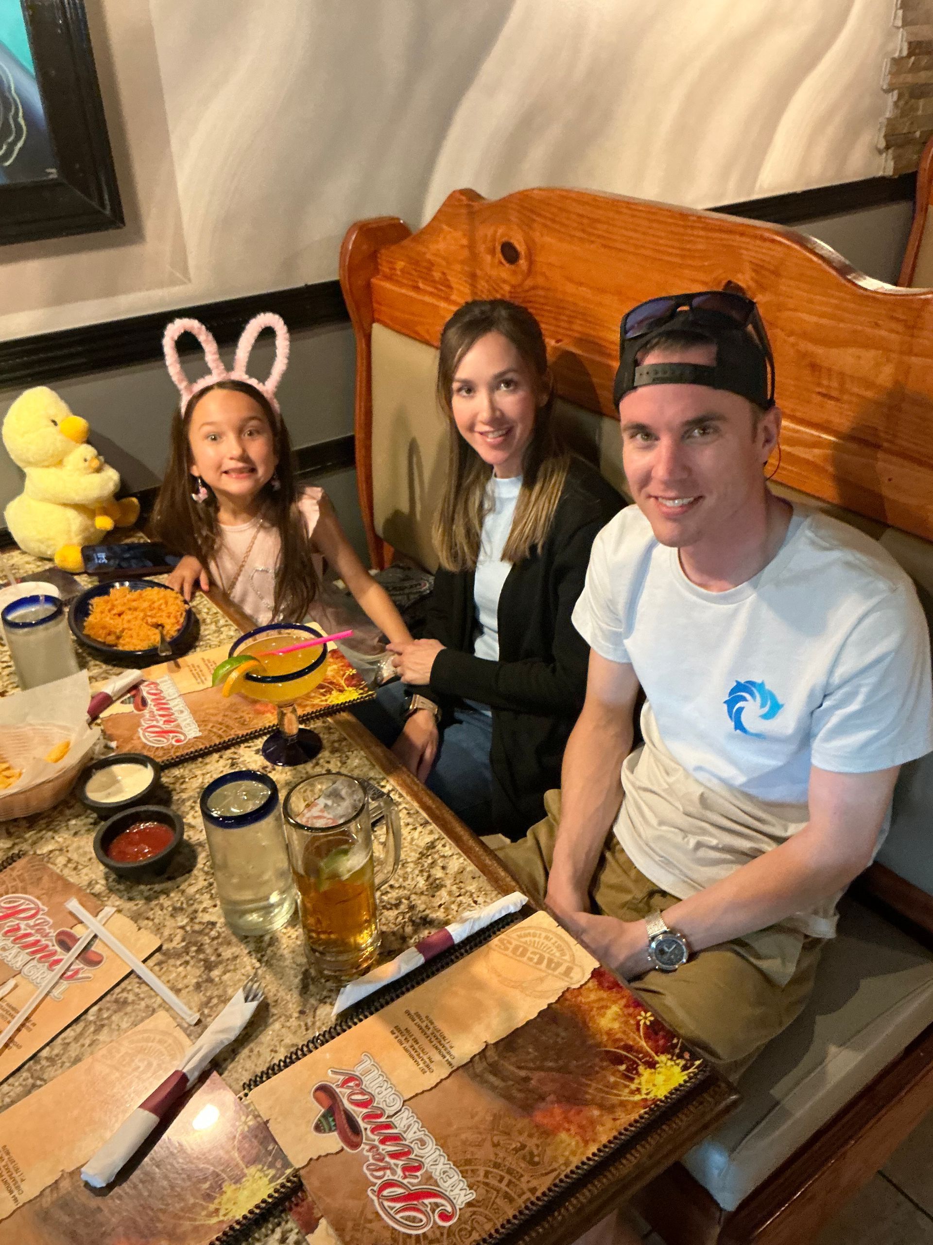Family at a restaurant table, smiling. A child wears bunny ears. Food and drinks are on the table.