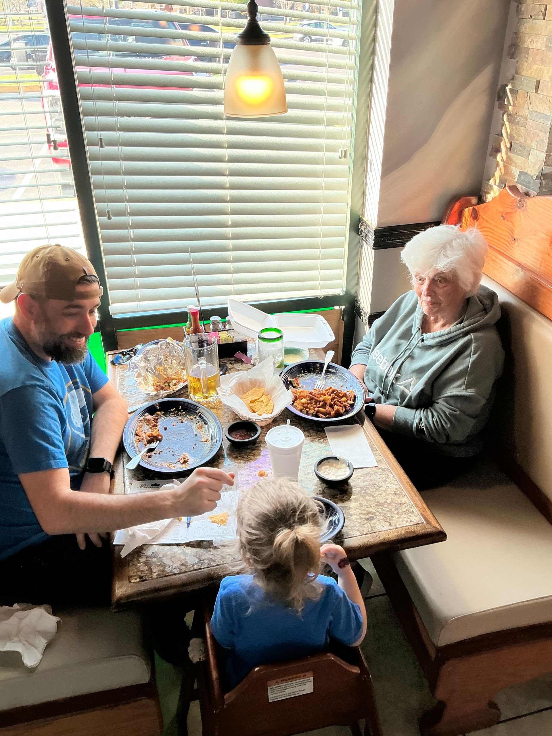Family eating at a restaurant table: man, child, and older woman seated, food on the table.
