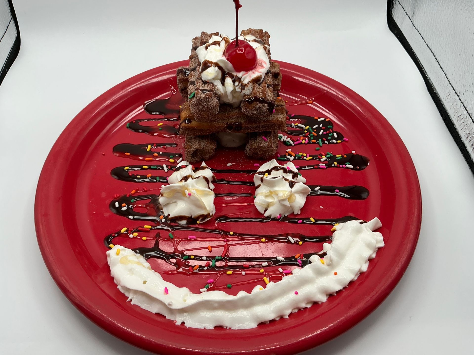 Chocolate dessert on red plate, decorated as a smiley face with whipped cream, chocolate syrup, sprinkles, and a cherry.