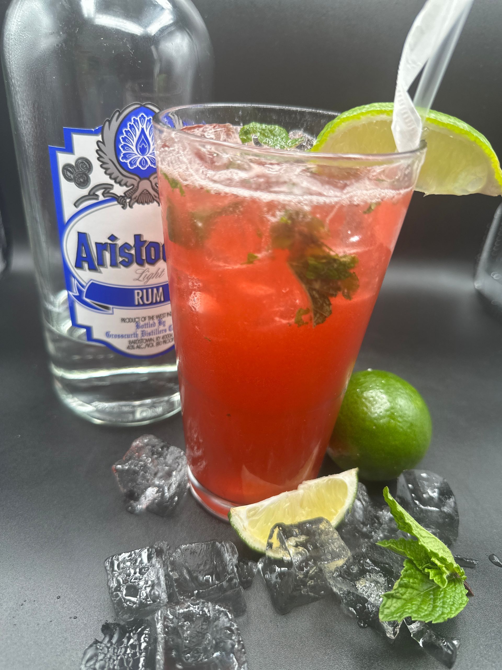 Red cocktail with ice, mint, and lime, next to a rum bottle.