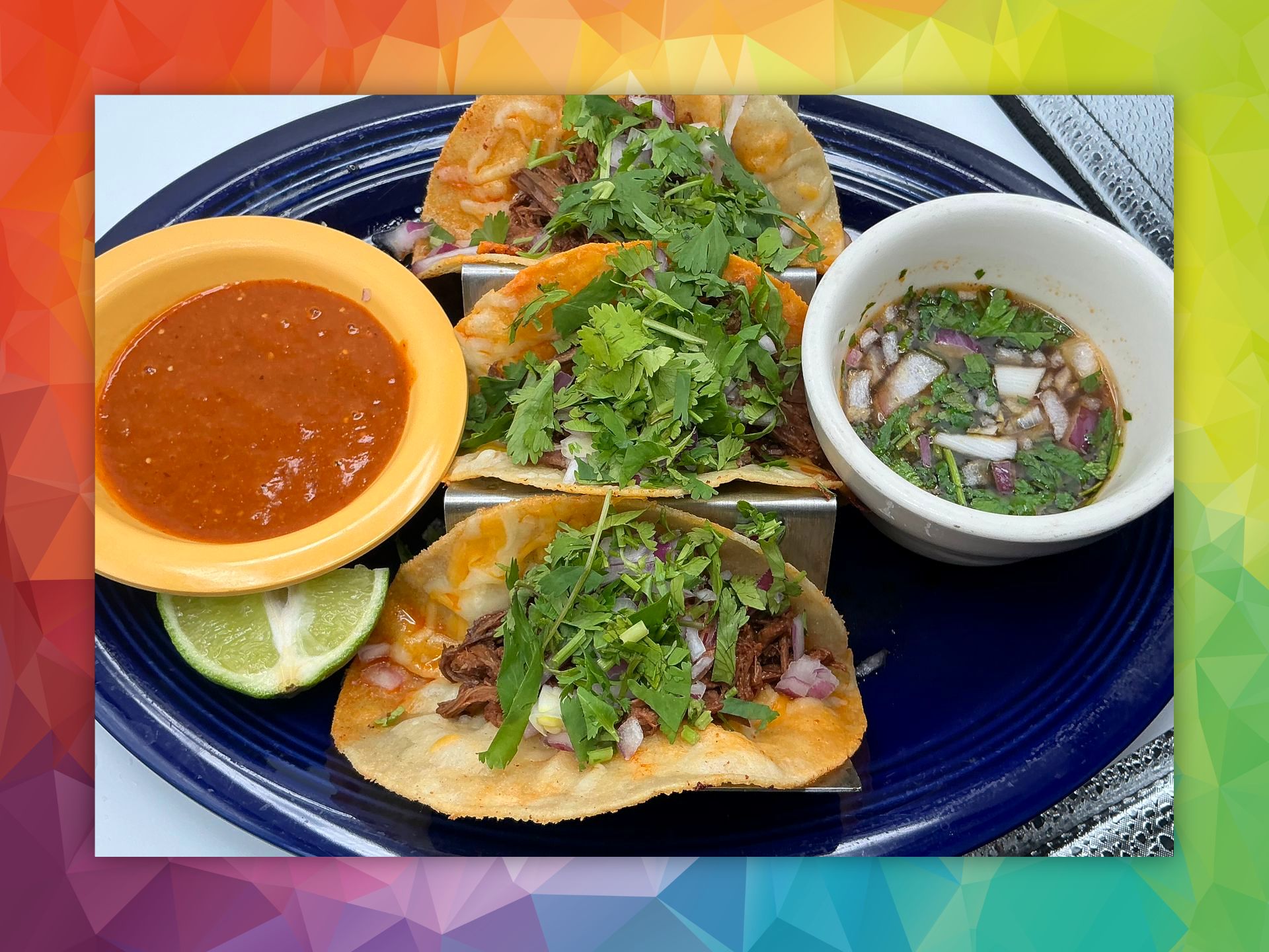Tacos on blue plate with salsa and dipping sauce, garnished with cilantro, and lime.