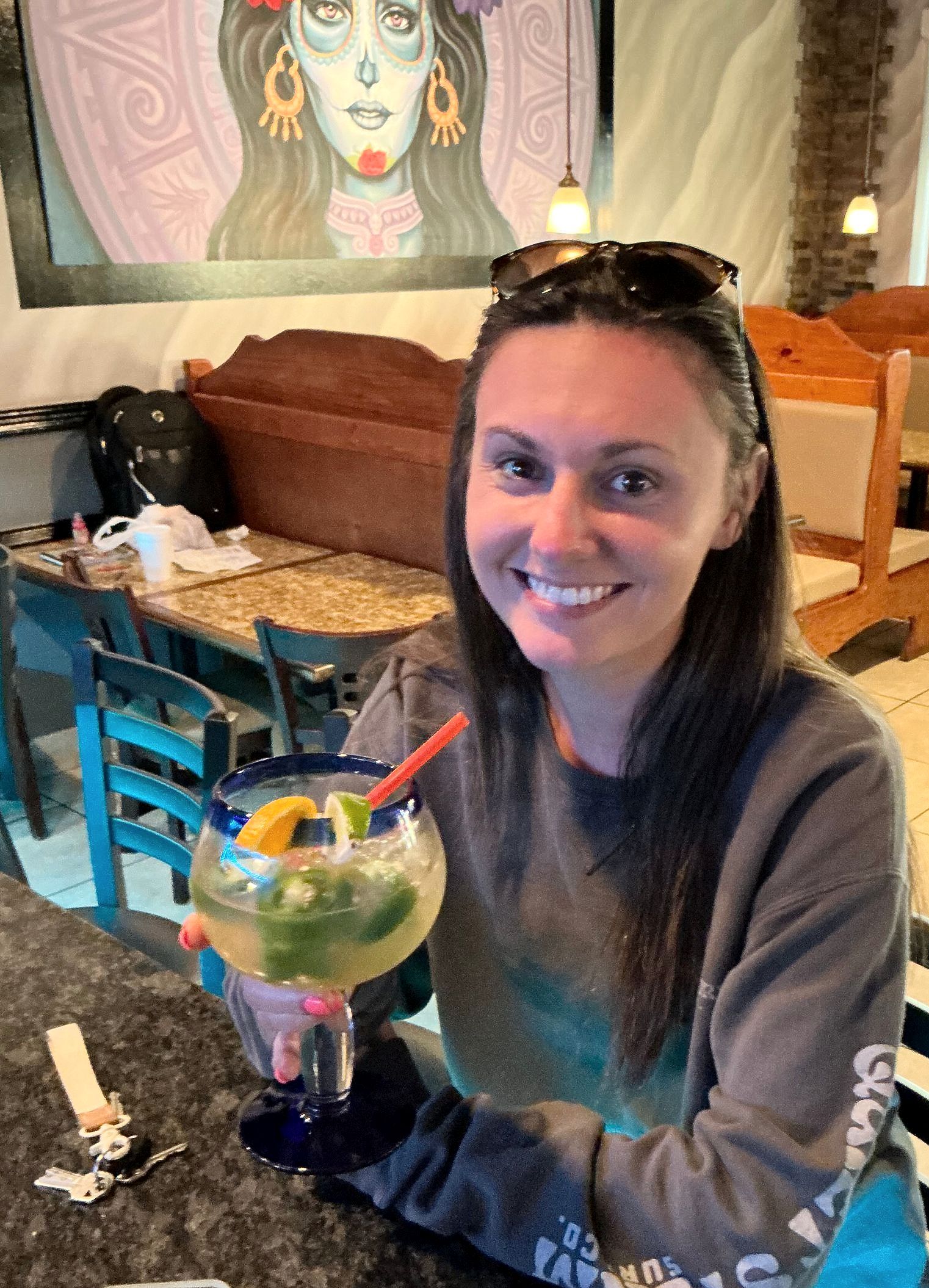 Woman smiling, holding a margarita with a bar and decorated restaurant in the background.