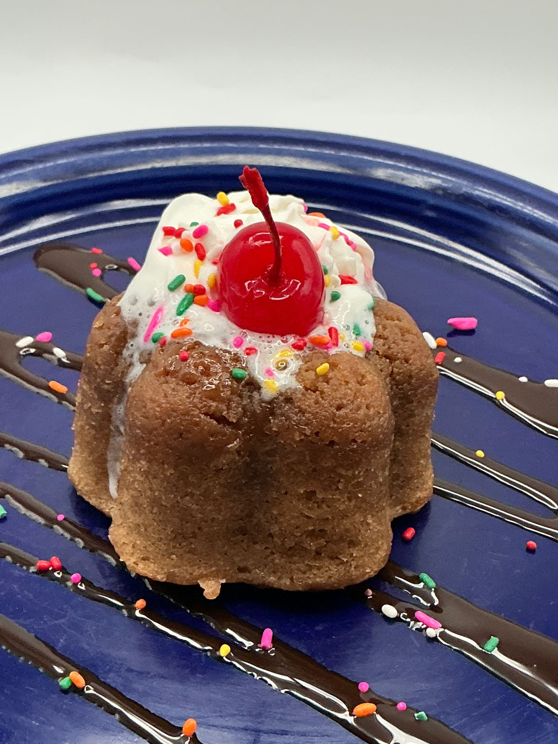 Small dessert cake with whipped cream, sprinkles, and a cherry on a blue plate drizzled with chocolate.