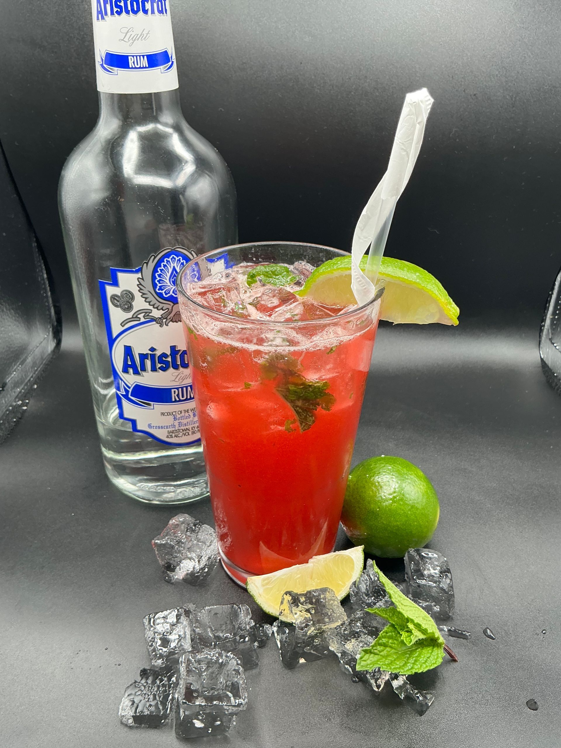 A vibrant red cocktail in a glass with a lime wedge and mint, next to a tequila bottle, limes, and ice on black.
