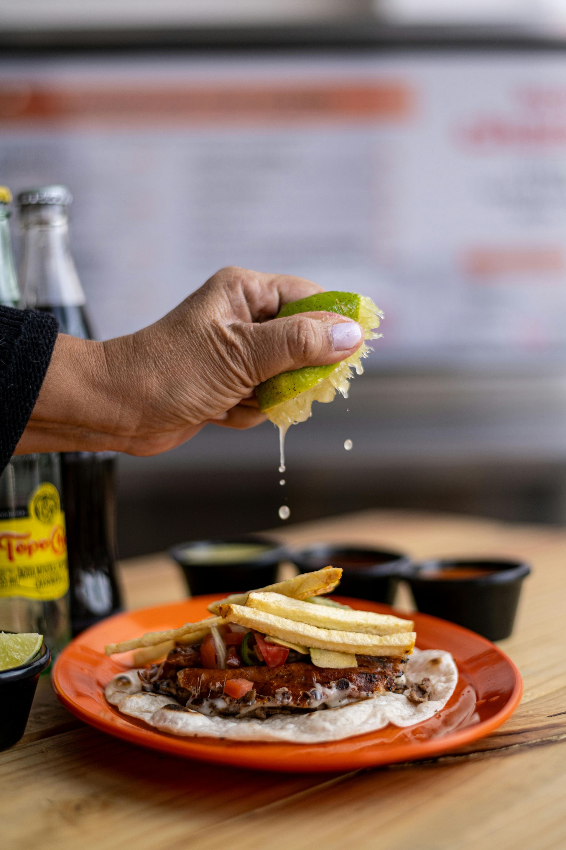 Hand squeezing lime juice onto a taco with toppings and french fries on an orange plate. Bottles of soda and sauces are nearby.