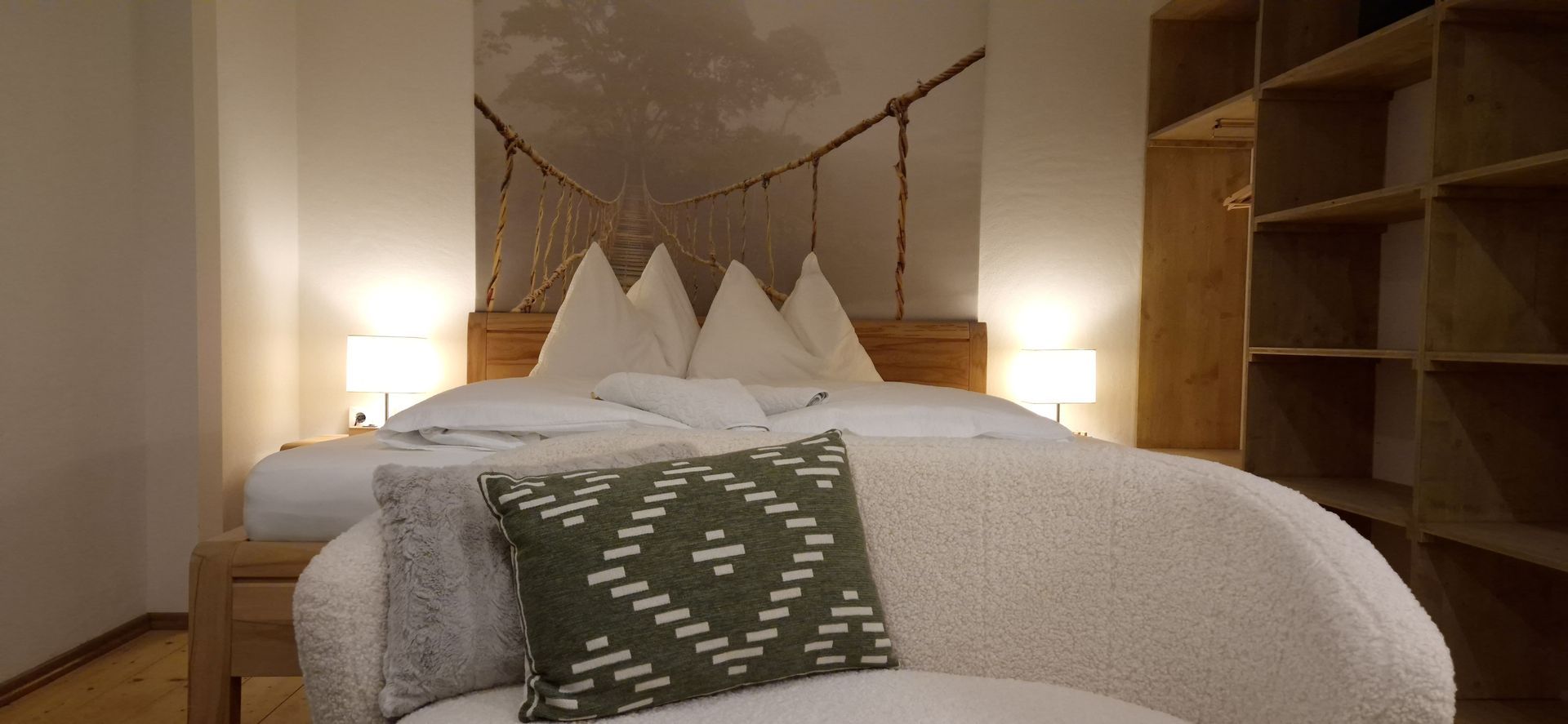 Gemütliches Bett mit weißen Kissen, gemusterter Tagesdecke, warmen Nachttischlampen und Holzregalen in einem Schlafzimmer.