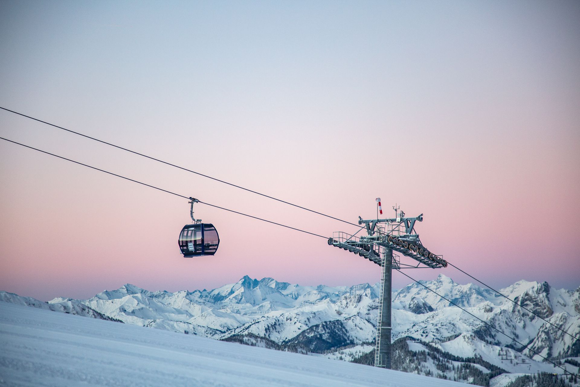 Skigondel über schneebedeckte Berge bei Sonnenuntergang, rosafarbener Himmel und Seilbahnturm im Vordergrund