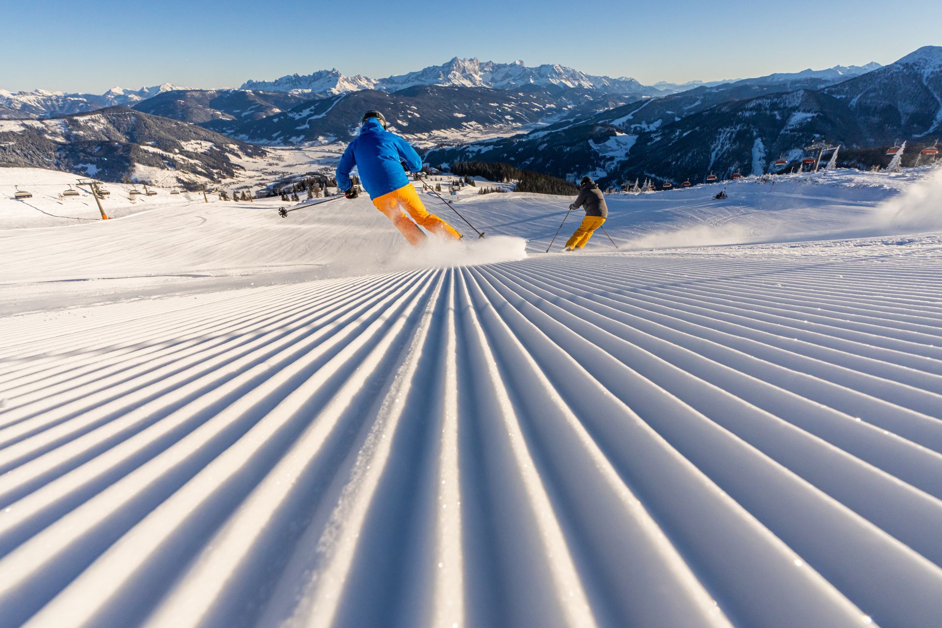 Skifahrer gleiten eine präparierte, schneebedeckte Piste hinab, im Hintergrund erheben sich Berggipfel.