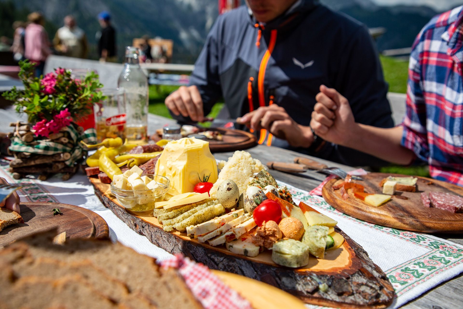 Picknicktisch im Freien mit Brot, Käse, Obst und im Hintergrund essende Menschen