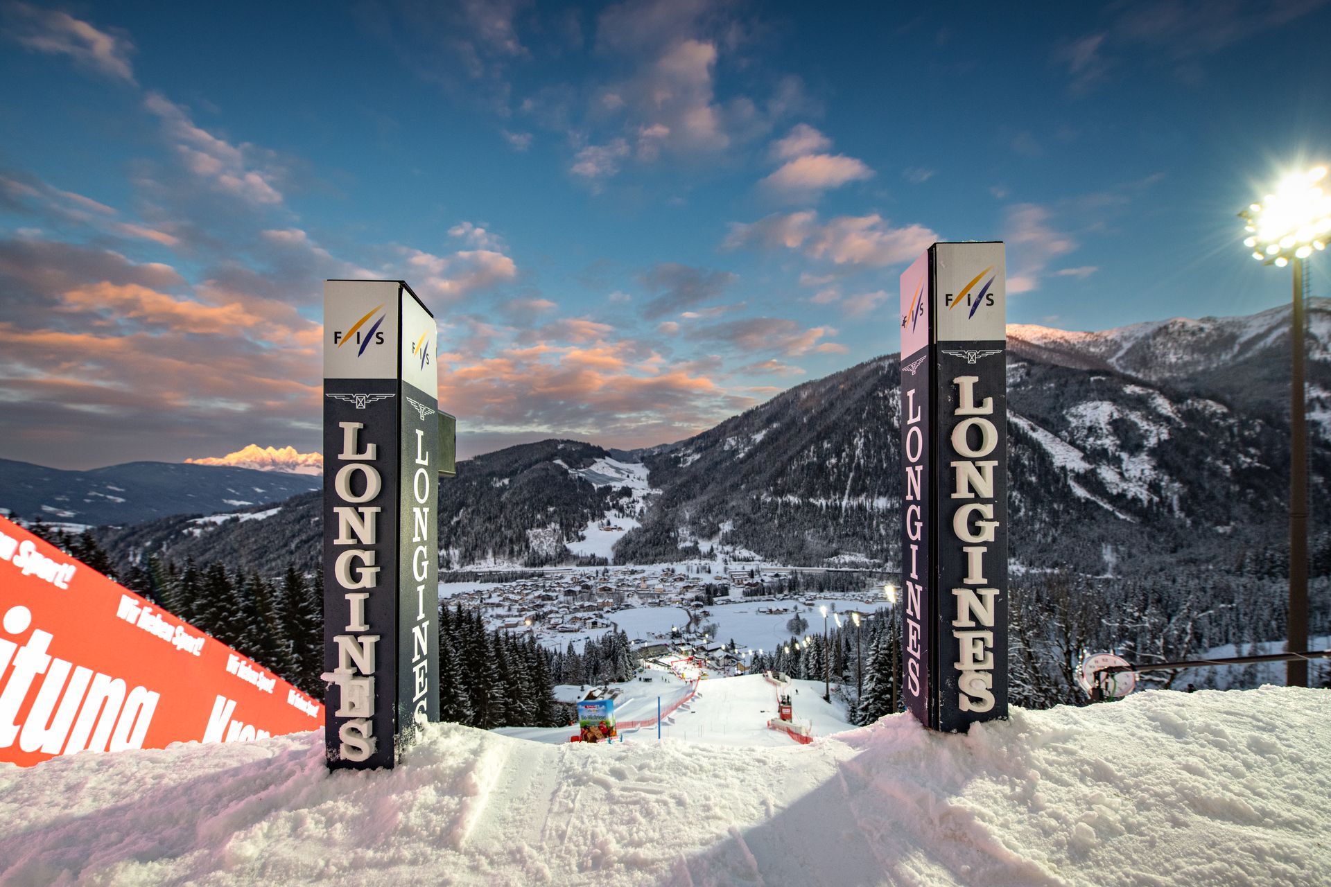 Verschneite Skipiste in der Abenddämmerung zwischen den Toren von Longines, mit Bergkulisse und rosablauem Himmel