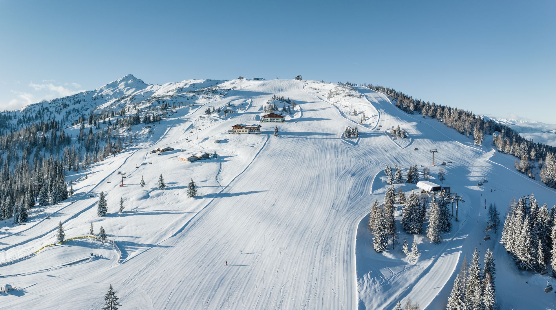 Schneebedeckte Skipiste an einem Berg unter klarem, blauem Himmel, mit Sesselliften und Kiefern.