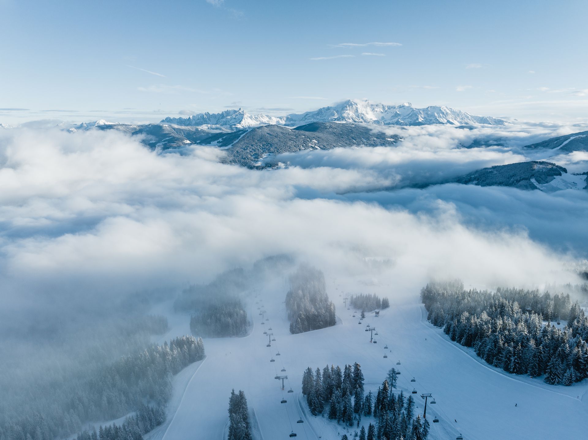 Verschneite Berglandschaft mit Wolken, bewaldeten Hängen und fernen Gipfeln unter klarem, blauem Himmel