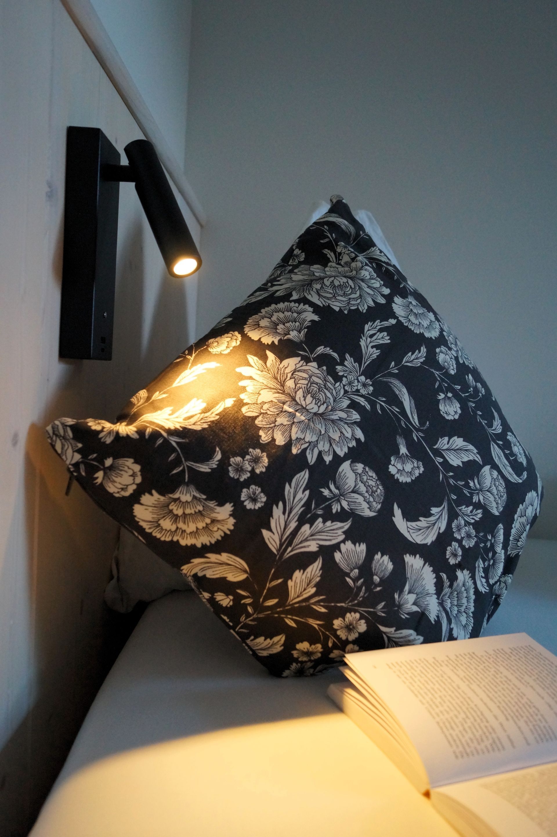 Eine Nachttischlampe beleuchtet ein schwarzes Kissen mit Blumenmuster neben einem offenen Buch auf einem weißen Bett.