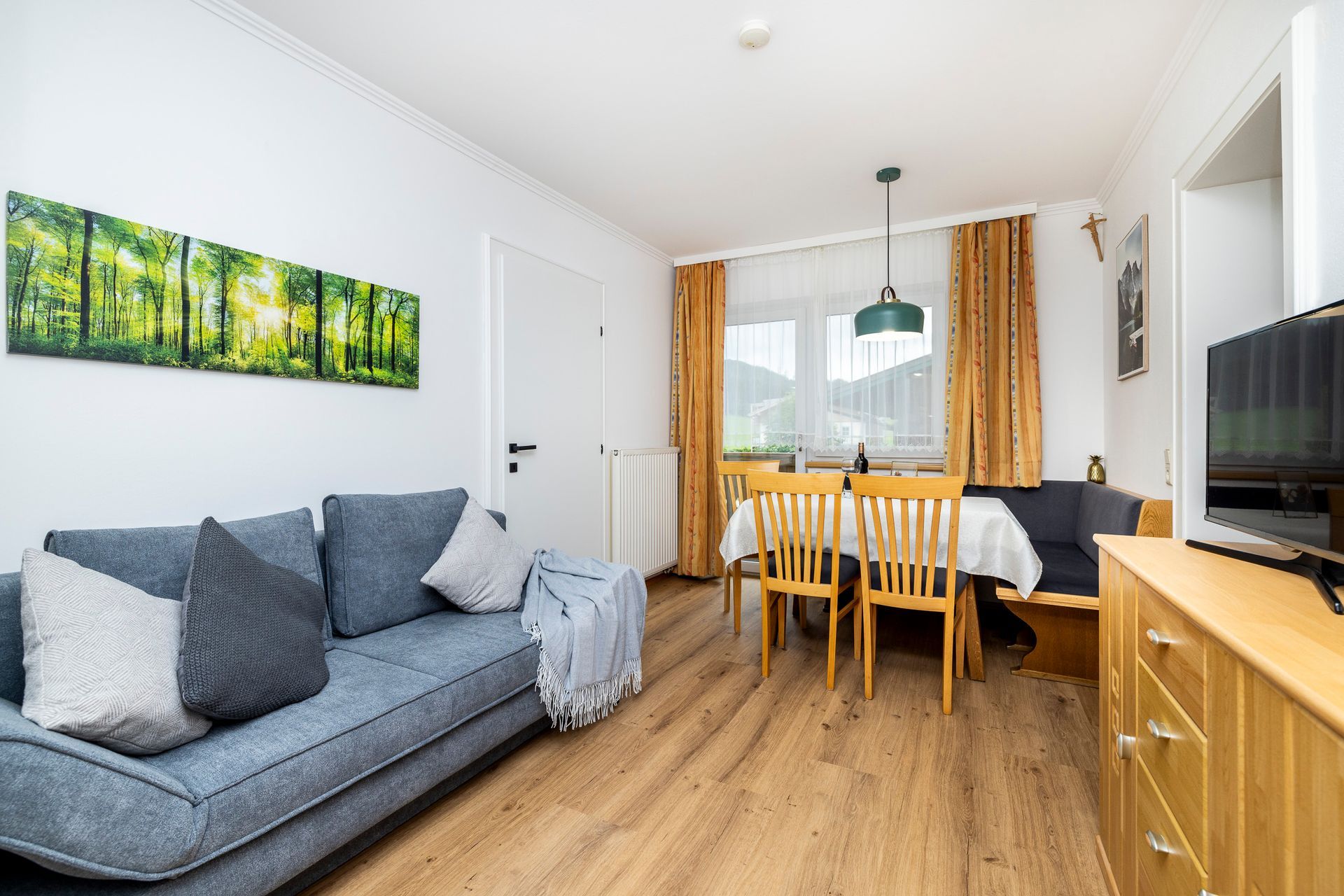 Helles Wohn-Esszimmer mit grauem Sofa, Holztisch, Fernseher und Wandbild mit Waldmotiven.
