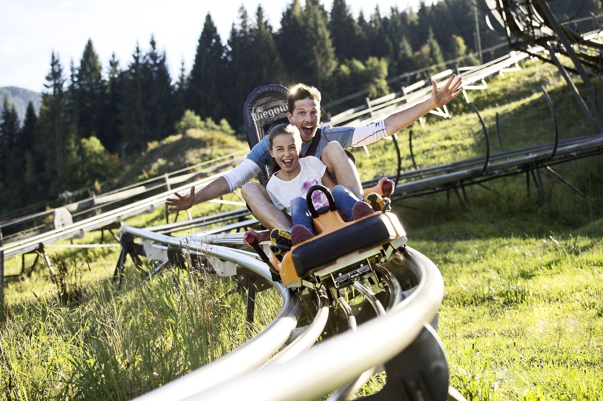 Zwei Personen fahren mit einer Bergachterbahn durch einen grasbewachsenen Hang, im Hintergrund sind Bäume zu sehen.