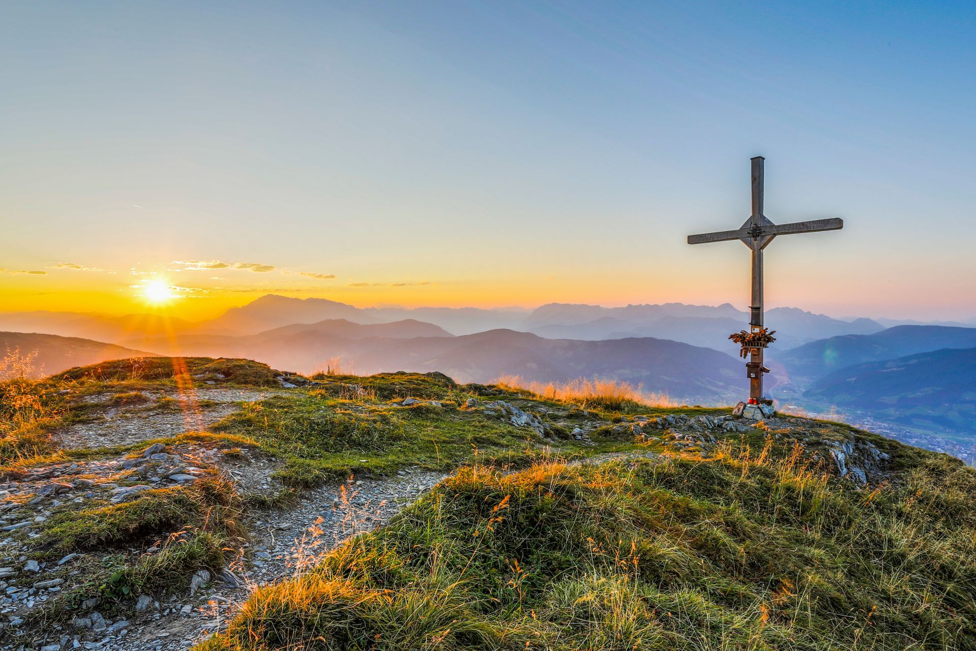 Sonnenaufgang über Berggipfeln mit einem Kreuz auf einem grasbewachsenen Hügel